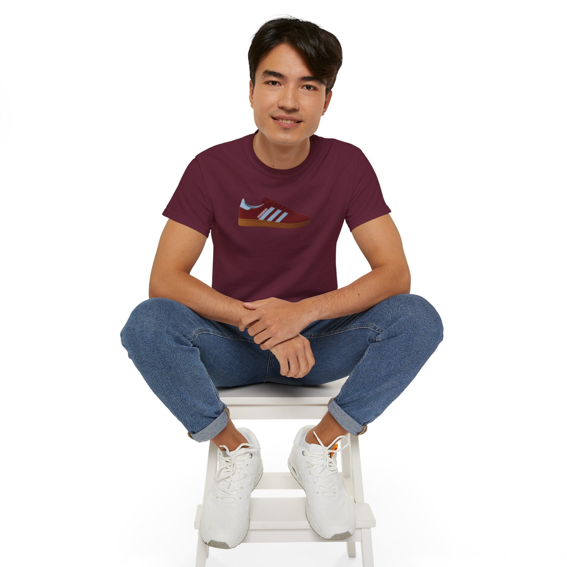 Young man sitting on white stool wearing maroon t-shirt with sneaker graphic and blue jeans