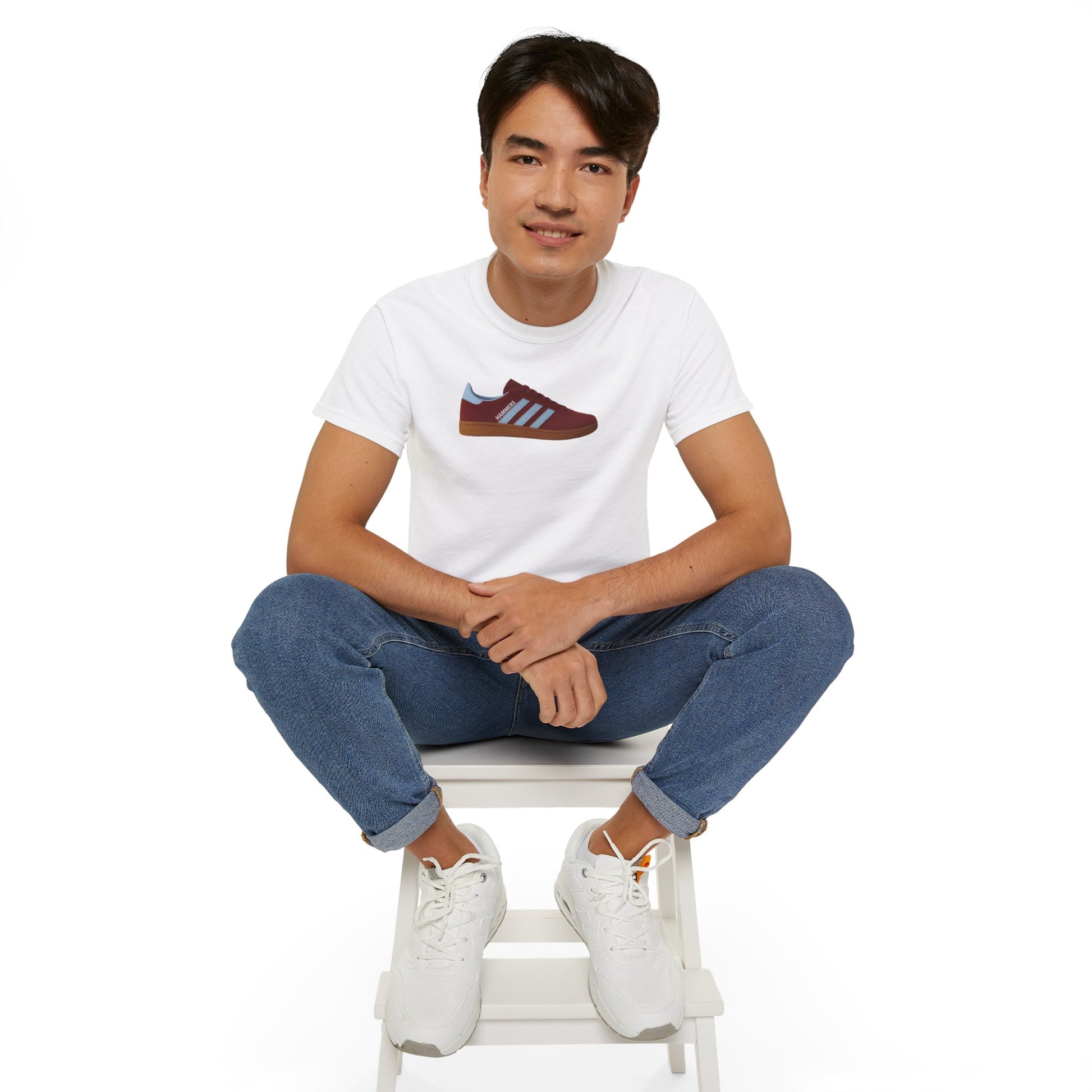 Young man wearing white graphic t-shirt with sneaker print, blue jeans, and white sneakers sitting on white stool
