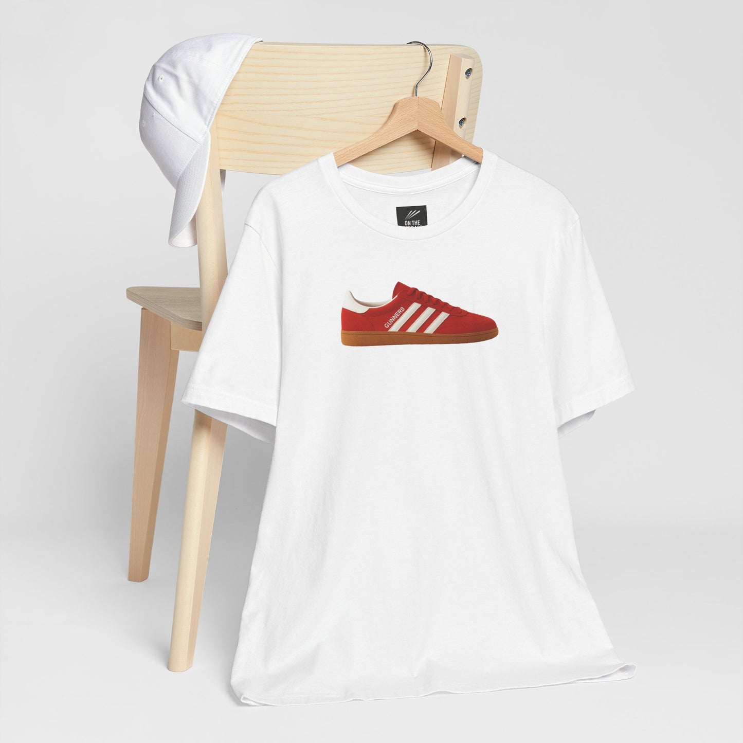 White t-shirt with red sneaker graphic and text hanging on wooden chair with white cap